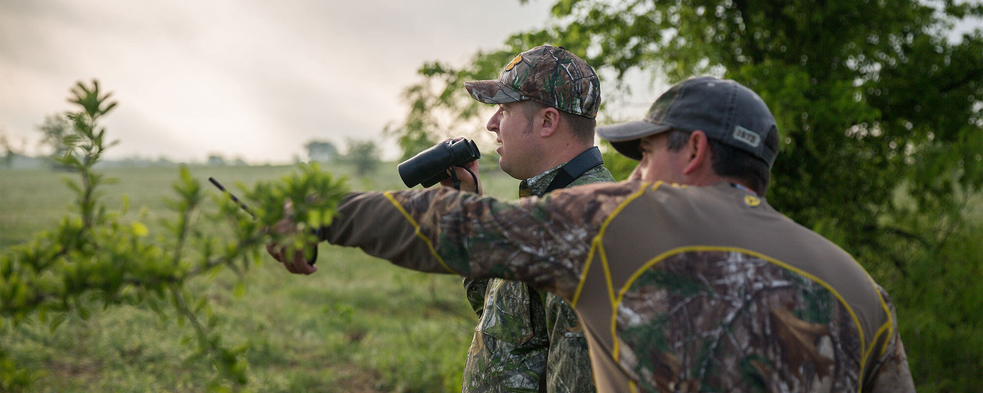 Two hunters standing on a hill looking across a field. One is holding binoculars and the other is pointing toward the field.