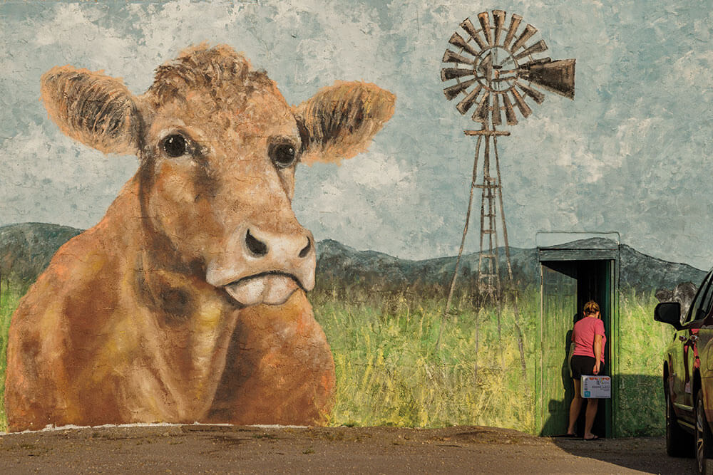 A lady walks into a building painted with a large mural of a cow standing in a pasture with mountains and a windmill in the background