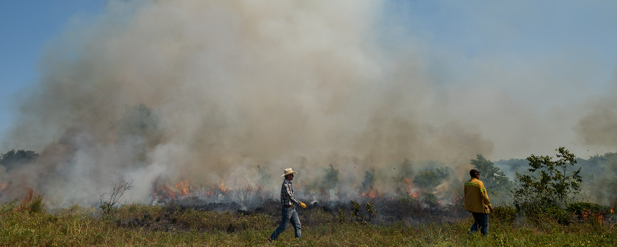 Ranchers conducting a prescribed burn look on as smoke rises from the fire.
