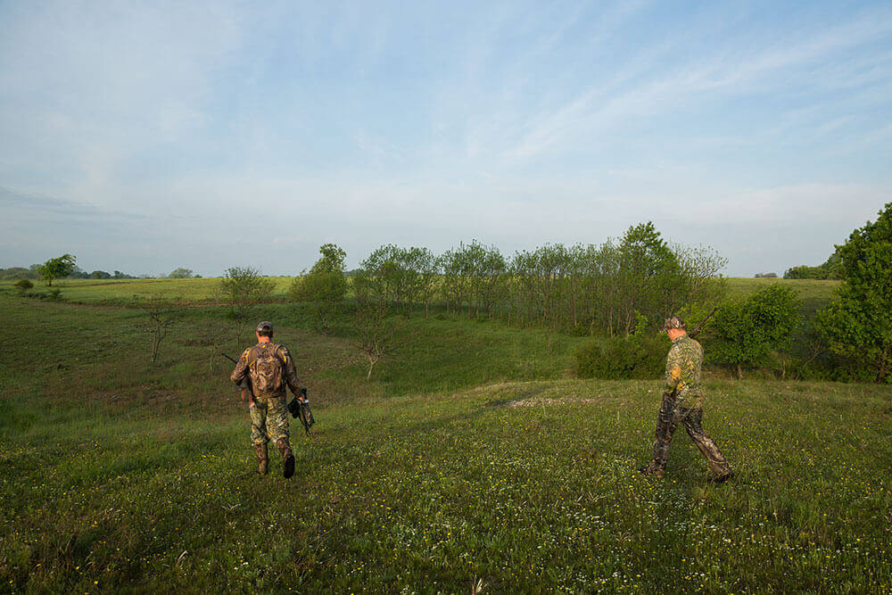 Two hunters with equipment walk across a field of hills rimmed with trees, in early morning sun
